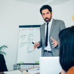 A man in a suit and tie delivers a presentation to an audience, gesturing confidently